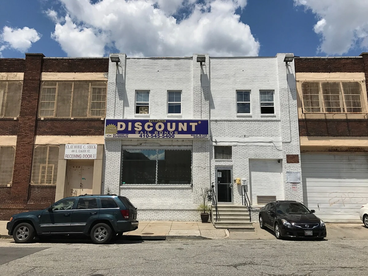 A white brick bail bonds office on East Eager Street in Baltimore with a blue sign that says 24 hour service, two cars parked at the curb