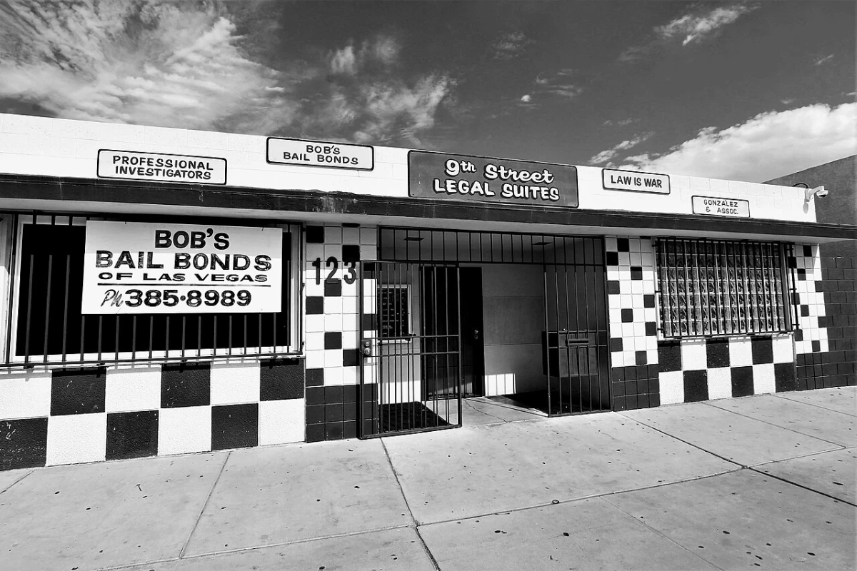 A black and white photo of a small bail bonds office in Las Vegas with a barred window, a large sign with a phone number and a checkered tile front