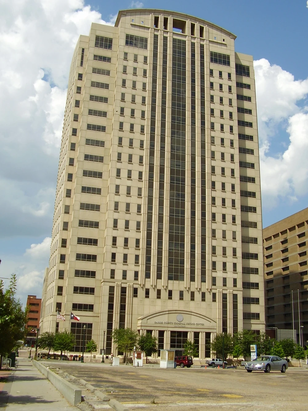 The Harris County Criminal Justice Center in downtown Houston, a tall tan stone tower with a curved top and an arched entrance, with a Texas flag and a United States flag out front